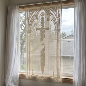 May include: A cream-coloured crochet window curtain featuring a central sword design framed by an intricate gothic arch pattern. The rectangular textile is suspended from a white rod and displays a detailed needlework motif of a sword blade.