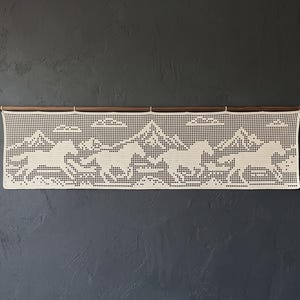 May include: A rectangular off-white crochet wall hanging featuring a repeating pattern of four horses running across a landscape with mountains and clouds in the background. The textile is suspended from a thin, horizontal wooden rod.