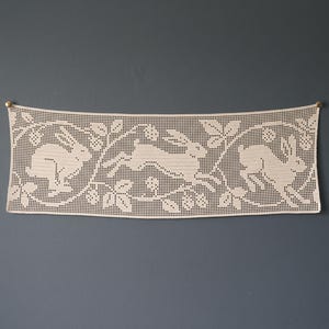 May include: A rectangular off-white crochet wall hanging featuring a repeating pattern of three leaping rabbits surrounded by leafy vines and small berries. The piece is secured at the top corners by two small gold-toned metal pins.