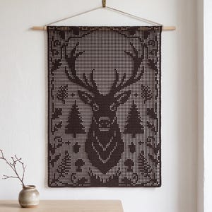 May include: A dark brown rectangular crochet wall hanging featuring a detailed deer head silhouette at the centre, surrounded by pine tree and foliage motifs. The piece is suspended from a light-coloured wooden dowel with a thin white cord.