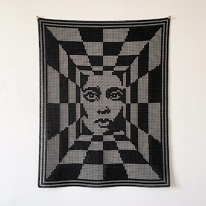 May include: A rectangular black and white crochet wall hanging featuring a central portrait of a face surrounded by a geometric, radiating checkerboard pattern that creates a deep, tunnel-like optical illusion effect across the entire piece.