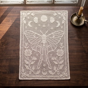 Peut inclure: Un napperon rectangulaire en crochet blanc cassé présentant un motif central de papillon de nuit, entouré de motifs floraux et d'un cycle de phases lunaires en haut. Le travail à l'aiguille affiche un motif en grille avec des étoiles.