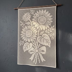 Könnte beinhalten: Ein cremefarbener, rechteckiger Wandbehang aus Häkelarbeit mit einem gitterbasierten Muster aus zwei Sonnenblumen mit Blättern und einer gebundenen Schleife am unteren Rand. Das Textil hängt an einem dunklen Holzstab mit einer dünnen Schnur.