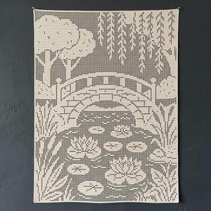 May include: A rectangular off-white crochet wall hanging featuring a detailed grid-style pattern of a stone bridge over a pond. The design includes water lilies, lily pads, tall reeds, and weeping willow trees, all rendered in a pixelated crochet stitch.