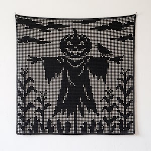 May include: A square black crochet wall hanging featuring a filet crochet design of a scarecrow with a jack-o'-lantern head standing in a cornfield under a cloudy sky with a bird perched on its shoulder, created with a grid-like openwork pattern.