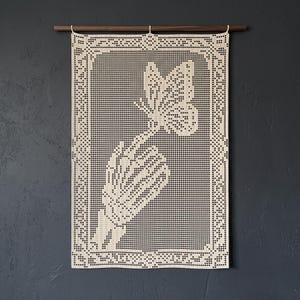 Peut inclure: Une tenture murale rectangulaire en crochet blanc cassé représentant une main de squelette tendant vers un papillon. La pièce est suspendue à une tige en bois foncé et présente un motif de bordure géométrique entourant le design central.