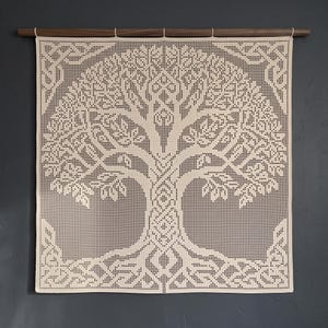 Op de afbeelding: Een vierkante, crèmekleurige gehaakte wandhanger met een gedetailleerd levensboomontwerp en ingewikkelde knooppatronen. Het textiel hangt aan een donkerbruine houten roede en vertoont een rasterachtige steekstructuur over het gehele oppervlak.