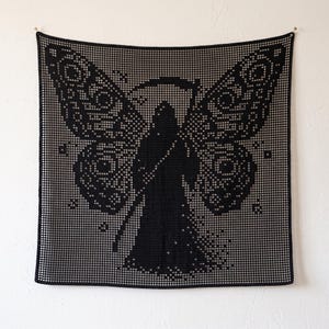 Könnte beinhalten: Ein quadratischer schwarzer Wandbehang aus Häkelarbeit mit einer zentralen Figur eines Sensenmanns mit Kapuze und Sense, flankiert von großen, komplexen Mottenflügeln. Das Design ist in einer gitterartigen Filethäkeltechnik gefertigt.