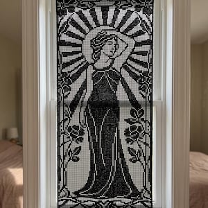 Puede incluir: Una cortina de ventana de ganchillo negro que presenta una figura detallada con un vestido largo, rodeada de motivos florales y un patrón de sol radiante. La pieza rectangular cuelga de una barra dorada, mostrando un intrincado trabajo de aguja.