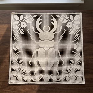 May include: A square off-white crochet doily featuring a central filet crochet design of a large beetle surrounded by floral motifs. The piece is crafted with a grid-like pattern of stitches, creating a detailed silhouette of the insect.