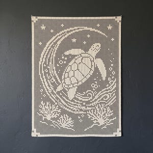 Op de afbeelding: Een rechthoekige off-white gehaakte wandhanger met een rasterpatroon van een zeeschildpad die in een grote halve maan zwemt, omringd door kleine sterren en koraalformaties aan de onderkant, gemaakt met fijn garen.