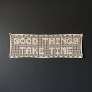May include: A rectangular off-white crochet wall hanging featuring a grid-like pattern. The piece displays the text GOOD THINGS TAKE TIME in a blocky, pixelated font style created by the negative space within the woven fiber construction.