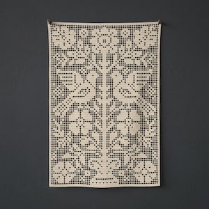 Op de afbeelding: Een rechthoekige beige gehaakte wandhanger met een rasterpatroon van twee vogels die naar elkaar toe kijken, omringd door bloem- en bladmotieven. Het handwerk creëert een symmetrisch ontwerp met open vierkante ruimtes door het hele werk.