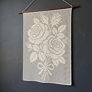 May include: A cream-coloured rectangular crochet wall hanging featuring a detailed floral pattern of roses and leaves tied with a bow. The textile is suspended from a dark brown wooden rod with a thin white string attached for hanging purposes.