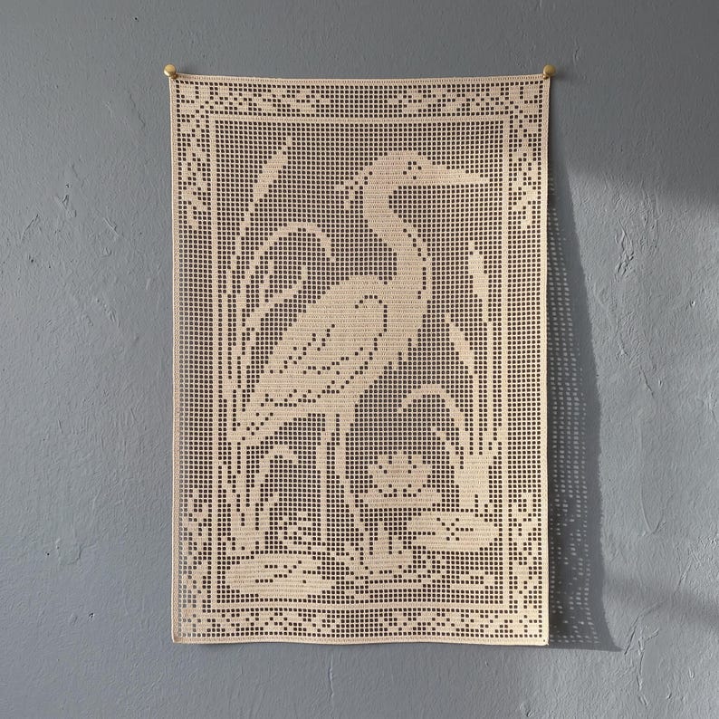 May include: A rectangular beige crochet wall hanging featuring a filet pattern of a heron standing among reeds and water lilies. The piece is bordered by a decorative geometric frame and is suspended from two small gold-colored metal pins.