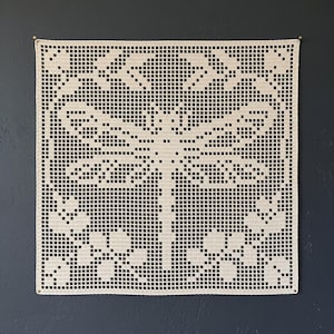 Puede incluir: Un tapete de ganchillo cuadrado en color blanco roto que presenta un motivo central de libélula con alas extendidas, rodeado de pequeños patrones florales. La pieza está hecha con técnica de ganchillo de red, creando un diseño geométrico calado.