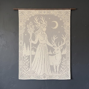 Könnte beinhalten: Ein cremefarbener Wandbehang aus Häkelarbeit mit einem gitterbasierten Muster einer Figur mit Geweih, die einen Stab hält und neben einem Hirsch steht. Das Design umfasst eine Mondsichel, Sterne und eine Waldlandschaft in einem Rahmen.