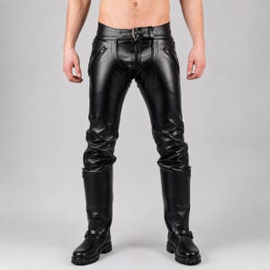 May include: Black leather trousers featuring a front zip fastening, a waist belt with a silver-toned metal buckle, and two diagonal zipped pockets on the hips. The trousers are paired with black leather knee-high boots with a low heel and side straps.