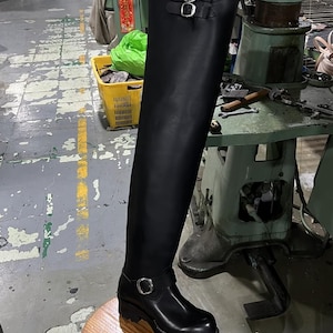 May include: A black leather over-the-knee boot featuring a rounded toe, a thick lug sole, and two silver-toned metal buckles. One buckle is positioned at the ankle, while the second buckle is located at the top outer edge of the boot shaft.