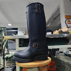 May include: A tall black leather boot featuring a rounded toe, a thick lugged rubber sole, and a low heel. The boot includes two adjustable straps with metal buckles at the top of the shaft and one adjustable strap with a metal buckle at the ankle.