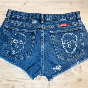May include: Dark blue denim shorts with white stitched skull designs on the back pockets. The shorts have a frayed hem and distressed details. A red Wrangler tag is visible.