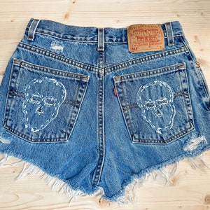 May include: Blue denim shorts with distressed details and white skull embroidery on the back pockets. The shorts feature a frayed hem and a Levi's brand tag.