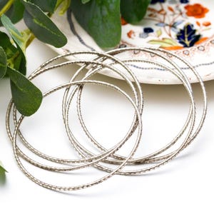 May include: A set of five thin, circular silver bangles featuring a textured, hammered surface. The bracelets are arranged in an overlapping, interlocking formation, creating a layered appearance with a metallic, reflective finish.