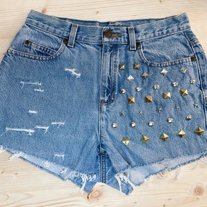 May include: Light blue denim shorts with distressed detailing and a raw hem. The right side is embellished with gold pyramid studs, clear rhinestones, and small star-shaped studs. A classic, casual style.
