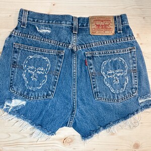 May include: Blue denim shorts with distressed details and frayed edges. The back pockets feature white stitched skull designs. A Levi Strauss & Co. label is visible. The shorts are a size W 28.