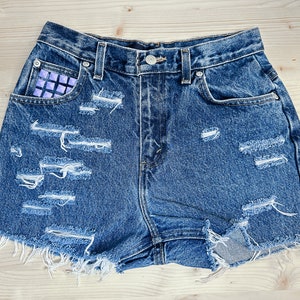 May include: Distressed denim shorts in a medium wash. The shorts feature multiple rips and tears, with frayed edges. The front pocket has a purple studded detail. The shorts have a classic five-pocket design and a button closure.