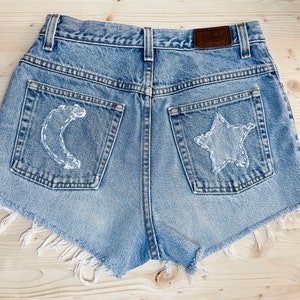 May include: Light blue denim shorts with a high waist and frayed edges. The back pockets feature white stitched designs of a crescent moon and a star. A brown leather patch is visible at the waistband.