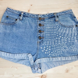 May include: Light blue denim shorts with a button fly and rolled cuffs. The right side features a white stitched design of lines, crosses, and circles. The shorts are laid flat on a light wood surface.