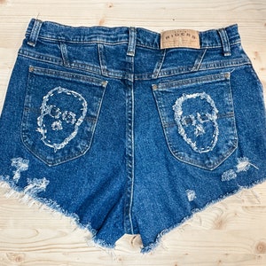 May include: Blue denim shorts with distressed edges and white skull embroidery on the back pockets. The shorts have a high waist and a "DENIM RIDERS" label. A unique, edgy fashion item.