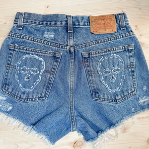 May include: Blue denim shorts with distressed details and white stitched skull designs on the back pockets. The shorts have a raw hem and a Levi's tag.