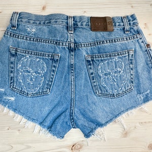 May include: Light blue denim shorts with distressed details and frayed edges. The back pockets feature white stitched skull designs. A brown leather patch with the text "Joans SL" is on the waistband.