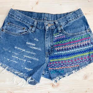 May include: Blue denim shorts with distressed detailing and colorful embroidered accents. The shorts feature a frayed hem and a decorative pattern of horizontal lines, triangles, and zigzags in various colors.
