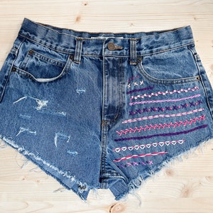 May include: Blue denim shorts with distressed details and colorful embroidered accents. The shorts feature a classic five-pocket design, a button closure, and frayed edges. The embroidery includes pink and purple stitching.