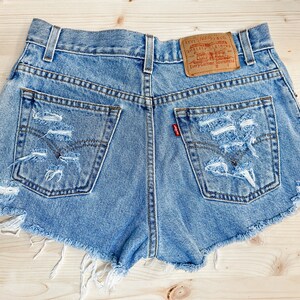 May include: Light blue denim shorts with distressed detailing, featuring ripped sections and frayed edges. The shorts have two back pockets, a red Levi's tag, and a brown leather label. The shorts are laid on a light wood surface.