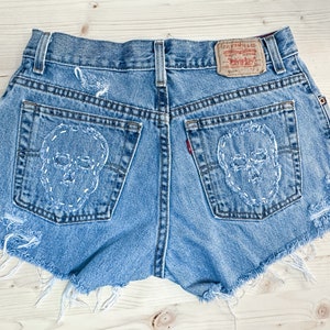 May include: Light blue denim shorts with distressed edges and white stitched skull designs on the back pockets. Features a Levi Strauss & Co. label. A unique, edgy fashion item.