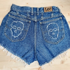 May include: Blue denim shorts with white skull embroidery on the back pockets. The shorts have a frayed hem and a leather "Lee" brand patch. A unique, edgy style.