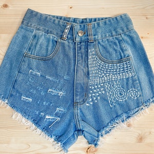 May include: Light blue denim shorts with distressed details and white decorative stitching. The shorts have a frayed hem, front pockets, and a button closure. The shorts are laid flat on a light wood surface.