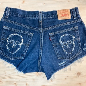 May include: Dark blue denim shorts with white skull embroidery on the back pockets. The shorts have a frayed hem and a Levi's brand tag. The shorts are a vintage style.