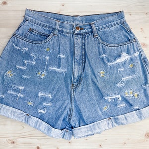 May include: Light blue denim shorts with distressed detailing and rolled-up cuffs. The shorts are adorned with embroidered yellow and white daisy flowers. A casual, summery style.