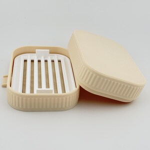 May include: A rectangular beige soap dish with a removable white slatted insert. The exterior of the dish features a vertical ribbed texture along the sides, and the matching beige lid is positioned partially open next to the base.