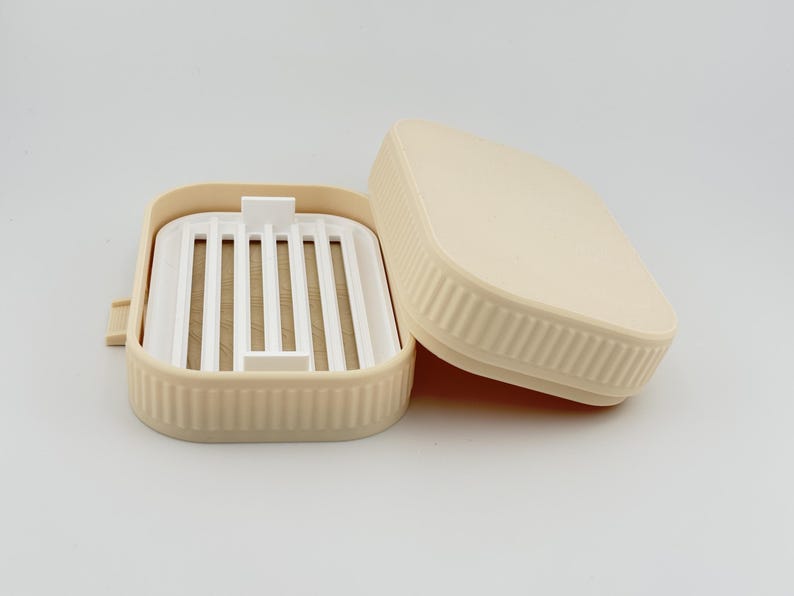 May include: A rectangular beige soap dish with a removable white slatted insert. The exterior of the dish features a vertical ribbed texture along the sides, and the matching beige lid is positioned partially open next to the base.