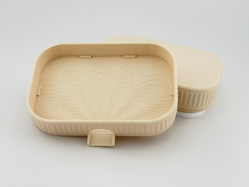 May include: A beige rectangular plastic container with a ribbed exterior and a matching lid. The interior base features a raised topographic line pattern. A small rectangular tab extends from the front edge of the container base.