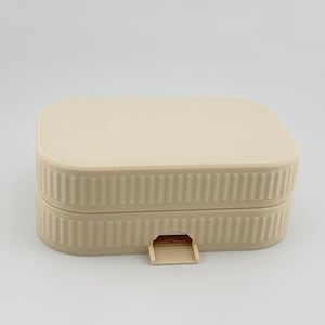 May include: A rectangular, light beige storage box with rounded corners and a ribbed, vertical texture along the sides. The container features a flat lid and a small, protruding tab at the center of the front edge for opening the lid.