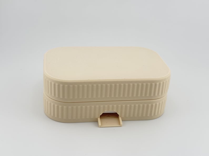 May include: A rectangular, light beige storage box with rounded corners and a ribbed, vertical texture along the sides. The container features a flat lid and a small, protruding tab at the center of the front edge for opening the lid.