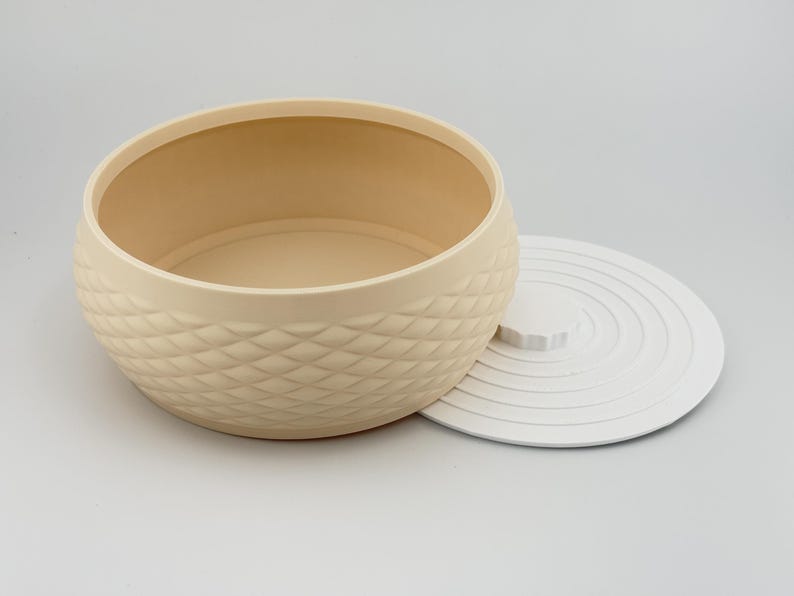 Puede incluir: Un recipiente de almacenamiento redondo de color beige con un exterior texturizado de patr&oacute;n de diamantes y una tapa blanca, plana y lisa, con anillos conc&eacute;ntricos y un peque&ntilde;o pomo central, presentado como un conjunto de dos piezas.