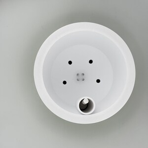 Looking straight down into the white soil insert, showing four small drainage holes, a tiny engraved part code in the centre, and the dark float bead sitting in the water gauge tube at the edge.